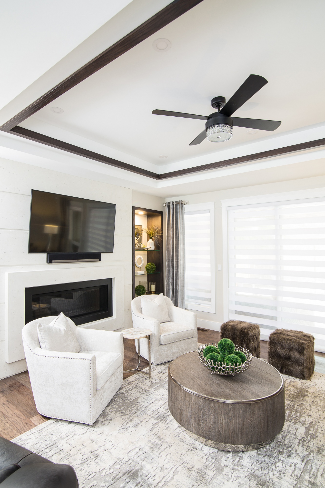 Modern living room with neutral tones, featuring a black-trimmed ceiling, sleek fireplace, white armchairs, round wooden coffee table with decorative bowl, and fur accent stools on a textured rug.