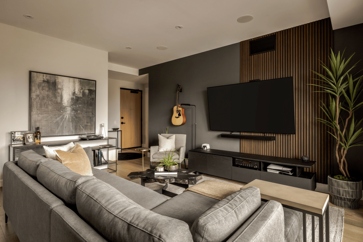 After photo of a transformed condo living room featuring a large grey sectional, mounted TV, warm wood slat wall, and modern accents including a guitar on display.