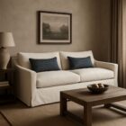 Cream slipcovered sofa with relaxed lines, two blue lumbar pillows, and tailored arms, shown in a styled neutral-toned living room and product view.