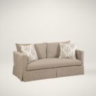 Neutral slipcovered sofa with tailored arms and damask accent pillows, featured in a traditional styled living room and studio background.