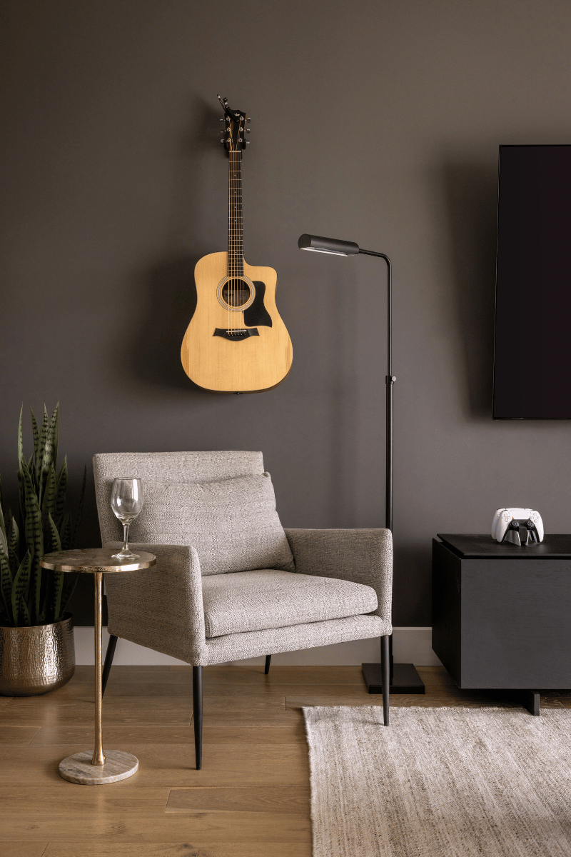 Minimalist reading nook with a grey armchair, floor lamp, and wall-mounted acoustic guitar, set against a dark accent wall.