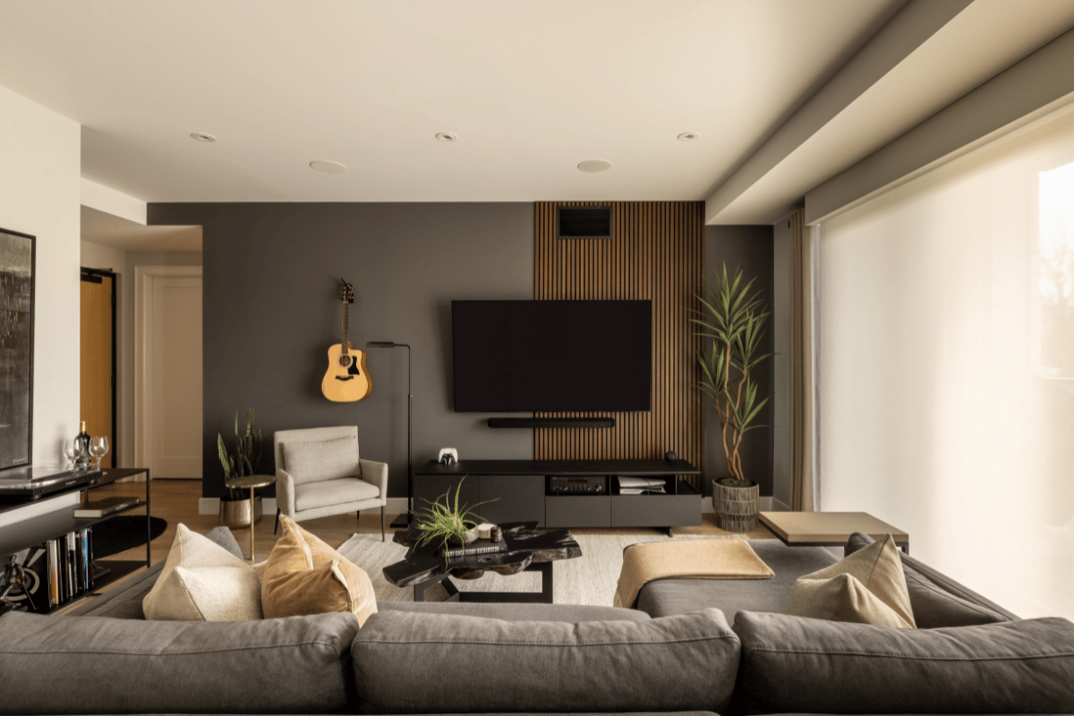 Modern living room featuring a TV wall with slatted wood panel detail, minimalist grey sectional, and natural light filtering through sheer window coverings.