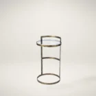 Small round accent table with an antique brass-finished frame and a clear glass top, styled beside a modern armchair in a neutral living space.