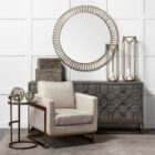 Small round accent table with an antique brass-finished frame and a clear glass top, styled beside a modern armchair in a neutral living space.