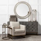 Small round accent table with an antique brass-finished frame and a clear glass top, styled beside a modern armchair in a neutral living space.