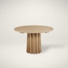 A round natural wood dining table with a sculptural, fluted pedestal base, styled on a soft neutral background.