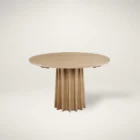 A round natural wood dining table with a sculptural, fluted pedestal base, styled on a soft neutral background.