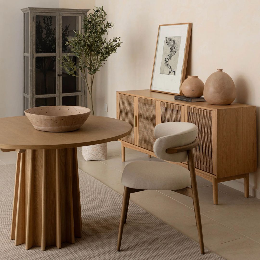 A round natural wood dining table with a sculptural, fluted pedestal base, styled on a soft neutral background.