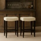 A backless upholstered saddle stool with cream fabric seat, dark espresso wood frame, and footrest support.