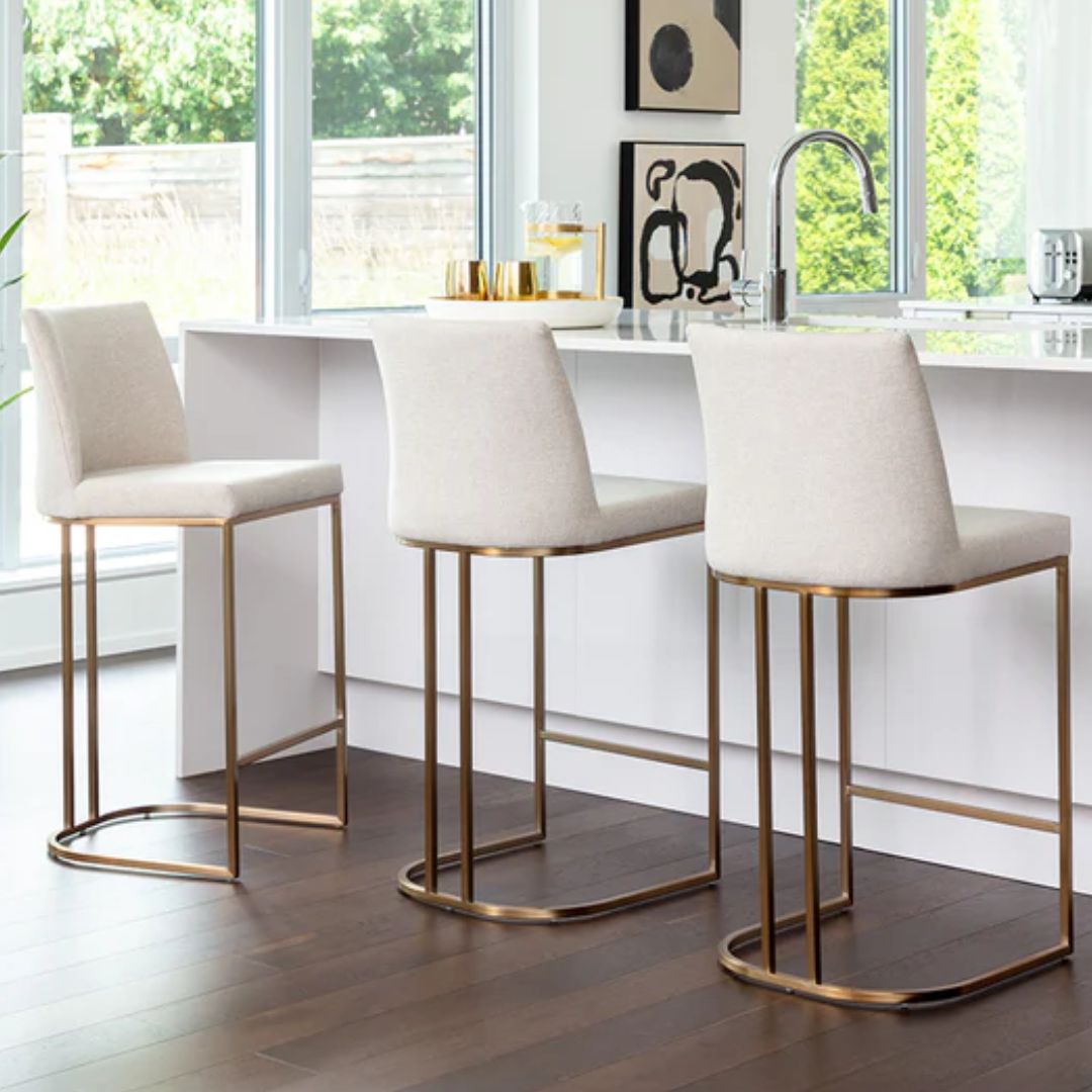A modern upholstered counter stool with a curved brass-finished metal base and light neutral fabric seat and back.