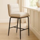 A modern upholstered counter stool with a soft cream fabric seat and backrest, wooden trim, and sleek black metal legs.