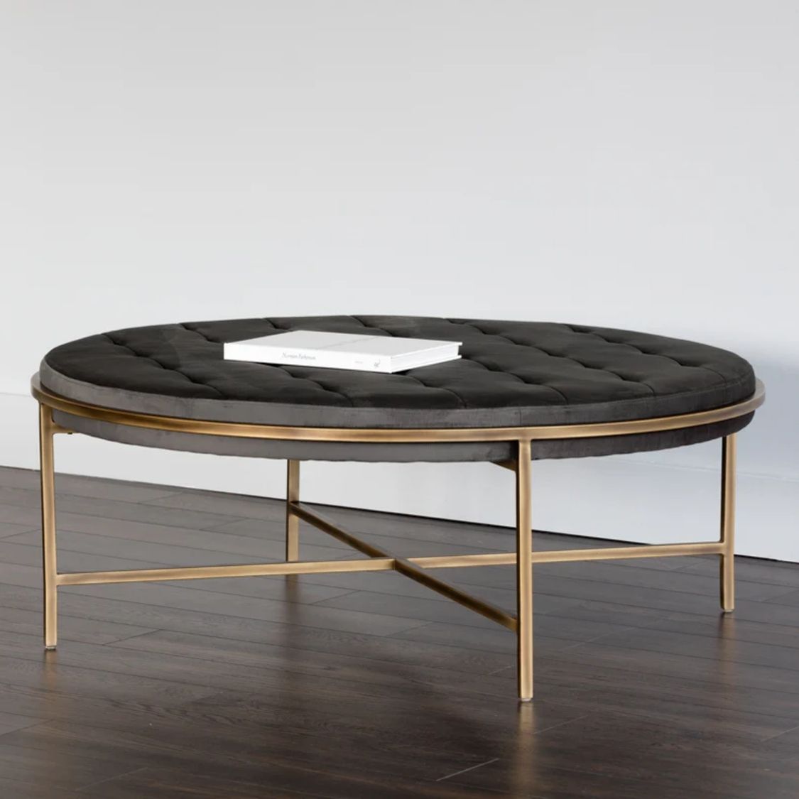 Round tufted ottoman with charcoal black upholstery and slender antique brass base frame.