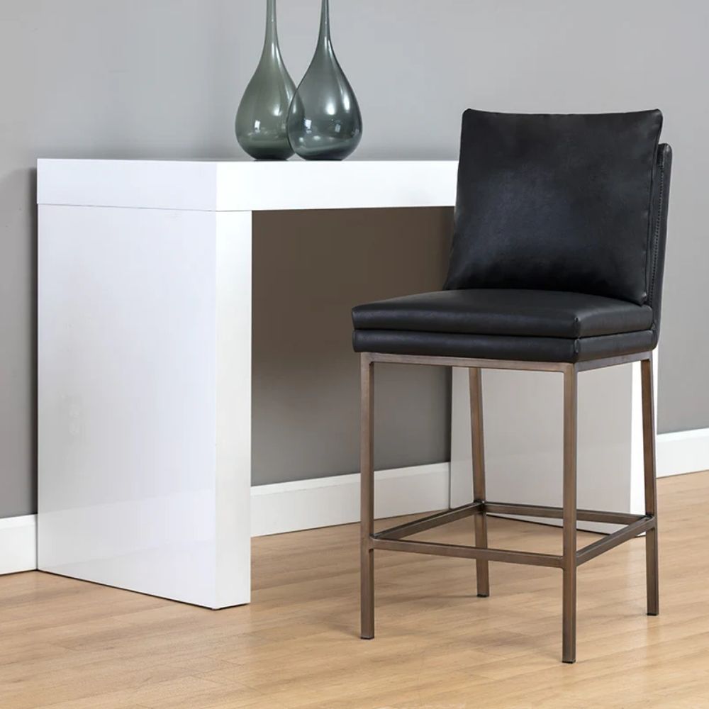 Paige Counter Stool featuring a black faux leather upholstered seat and loose-style back cushion, set on a brushed metal frame with footrest