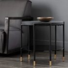 Round modern end table with black textured top, slim black legs, lower shelf, and brass-capped feet.
