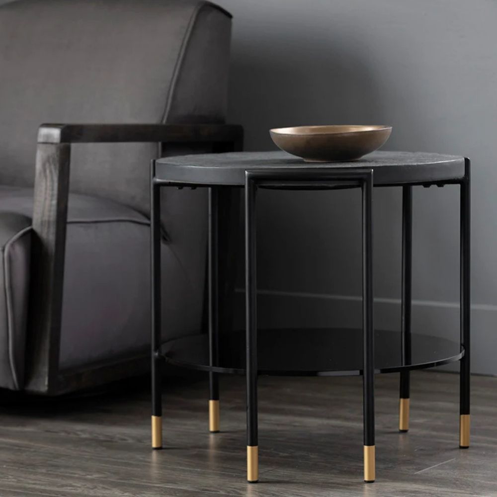 Round modern end table with black textured top, slim black legs, lower shelf, and brass-capped feet.
