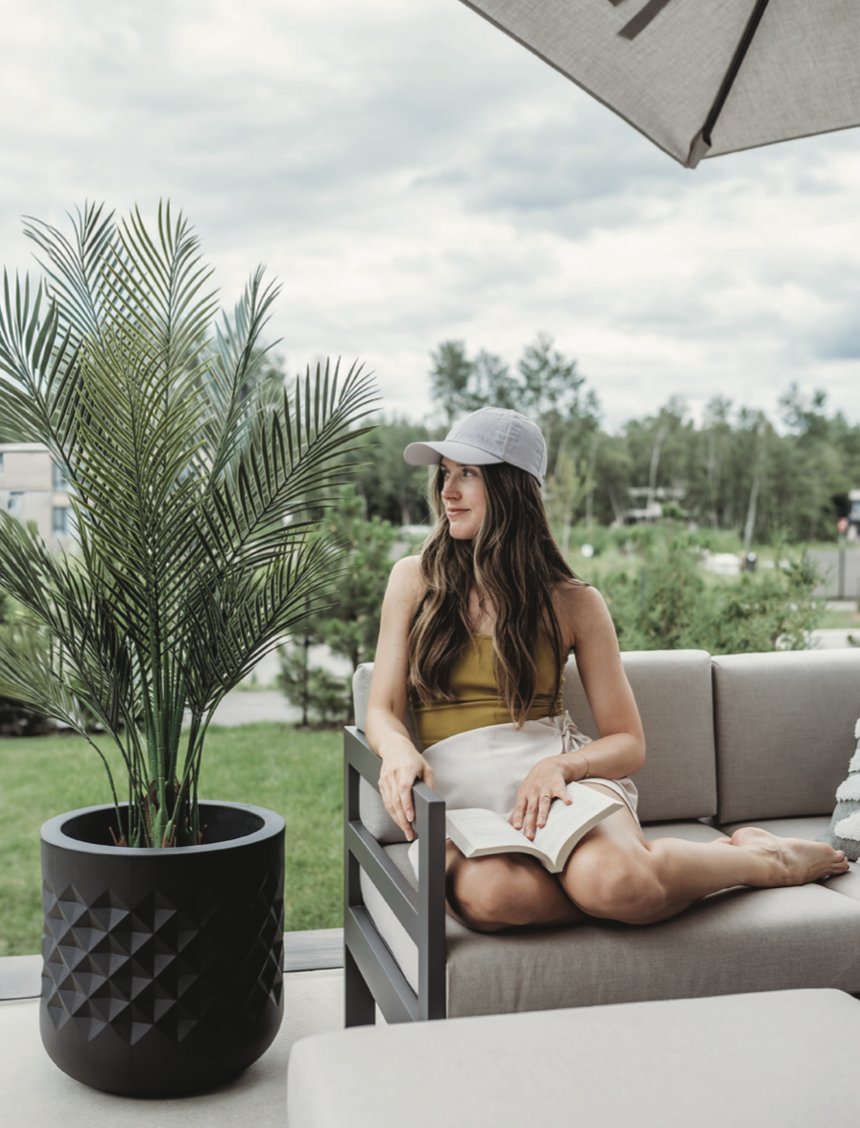 Faux palm tree in a matte black planter with geometric texture, next to a woman relaxing outdoors under an umbrella.