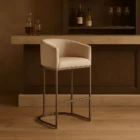 Front and side view of an elegant modern counter stool with a curved backrest, upholstered in a light neutral fabric with sleek metallic legs and a rounded base.