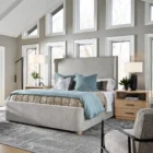 Upholstered bed with a high winged headboard in textured neutral fabric, rounded corners, and low wood feet, shown in both studio and lifestyle settings.