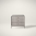 A modern three-drawer nightstand with soft, rounded corners and reeded drawer fronts, finished in a light neutral tone with gold hardware, styled beside a contemporary upholstered bed.