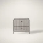 A modern three-drawer nightstand with soft, rounded corners and reeded drawer fronts, finished in a light neutral tone with gold hardware, styled beside a contemporary upholstered bed.
