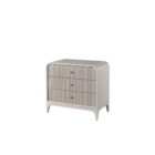 A modern three-drawer nightstand with soft, rounded corners and reeded drawer fronts, finished in a light neutral tone with gold hardware, styled beside a contemporary upholstered bed.