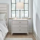 A modern three-drawer nightstand with soft, rounded corners and reeded drawer fronts, finished in a light neutral tone with gold hardware, styled beside a contemporary upholstered bed.