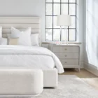 A modern three-drawer nightstand with soft, rounded corners and reeded drawer fronts, finished in a light neutral tone with gold hardware, styled beside a contemporary upholstered bed.