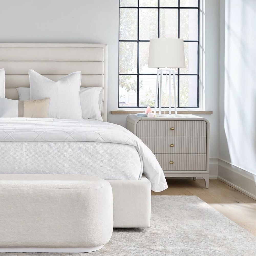 A modern three-drawer nightstand with soft, rounded corners and reeded drawer fronts, finished in a light neutral tone with gold hardware, styled beside a contemporary upholstered bed.