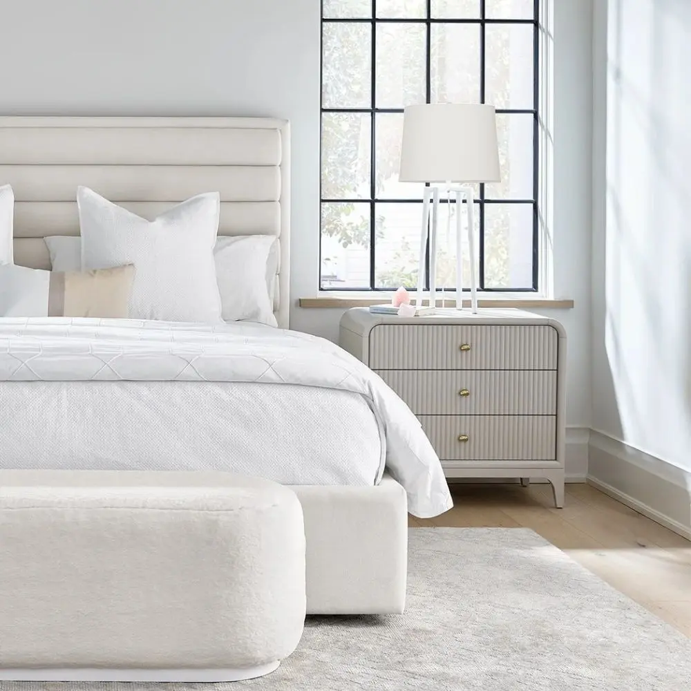 A modern three-drawer nightstand with soft, rounded corners and reeded drawer fronts, finished in a light neutral tone with gold hardware, styled beside a contemporary upholstered bed.