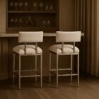 A modern counter stool featuring a soft upholstered back and seat in a neutral woven fabric, supported by a sleek polished metal frame with a footrest.
