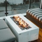 A sleek rectangular Dekko Avera 48 firepit in a minimalist concrete finish, showcasing a linear flame bed with lava rock fill.