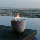 A modern Dekko Element firepit with barrelled concrete walls and a gently cupped-shaped body, filled with smooth black lava rock and warm flames.