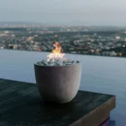 A modern Dekko Element firepit with barrelled concrete walls and a gently cupped-shaped body, filled with smooth black lava rock and warm flames.