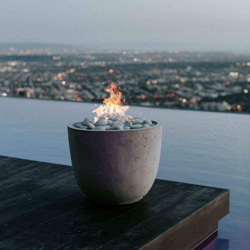 A modern Dekko Element firepit with barrelled concrete walls and a gently cupped-shaped body, filled with smooth black lava rock and warm flames.