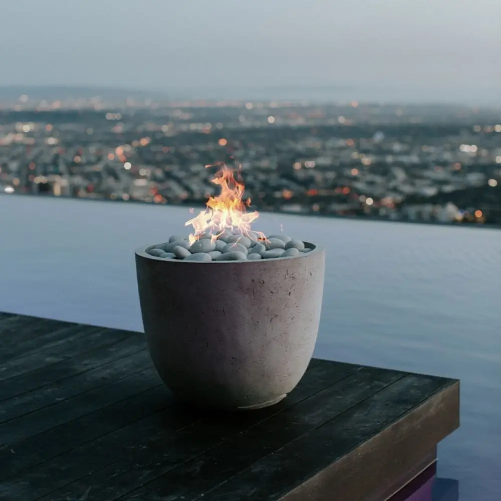 A modern Dekko Element firepit with barrelled concrete walls and a gently cupped-shaped body, filled with smooth black lava rock and warm flames.