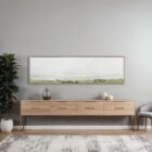A panoramic landscape wall art piece featuring soft textured strokes of green fields beneath a wide expanse of pale sky, framed in a slim border.