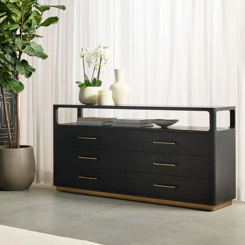 A modern black six-drawer dresser with slim brass handles, open top shelf, and a brass base accent.