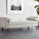 Contemporary upholstered bench with tufted seat, bolster cushions, and wooden legs in soft light fabric.