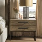 A modern nightstand in a light natural wood finish with two drawers, oversized black metal handles, and an open metal base frame.