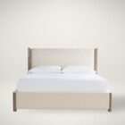 A contemporary upholstered bed in soft beige fabric with a tall padded headboard and natural wood trim accents on the corners and legs.
