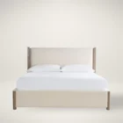 A contemporary upholstered bed in soft beige fabric with a tall padded headboard and natural wood trim accents on the corners and legs.