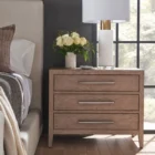 A three-drawer nightstand in a light natural wood finish with sleek horizontal metal handles and tapered legs.