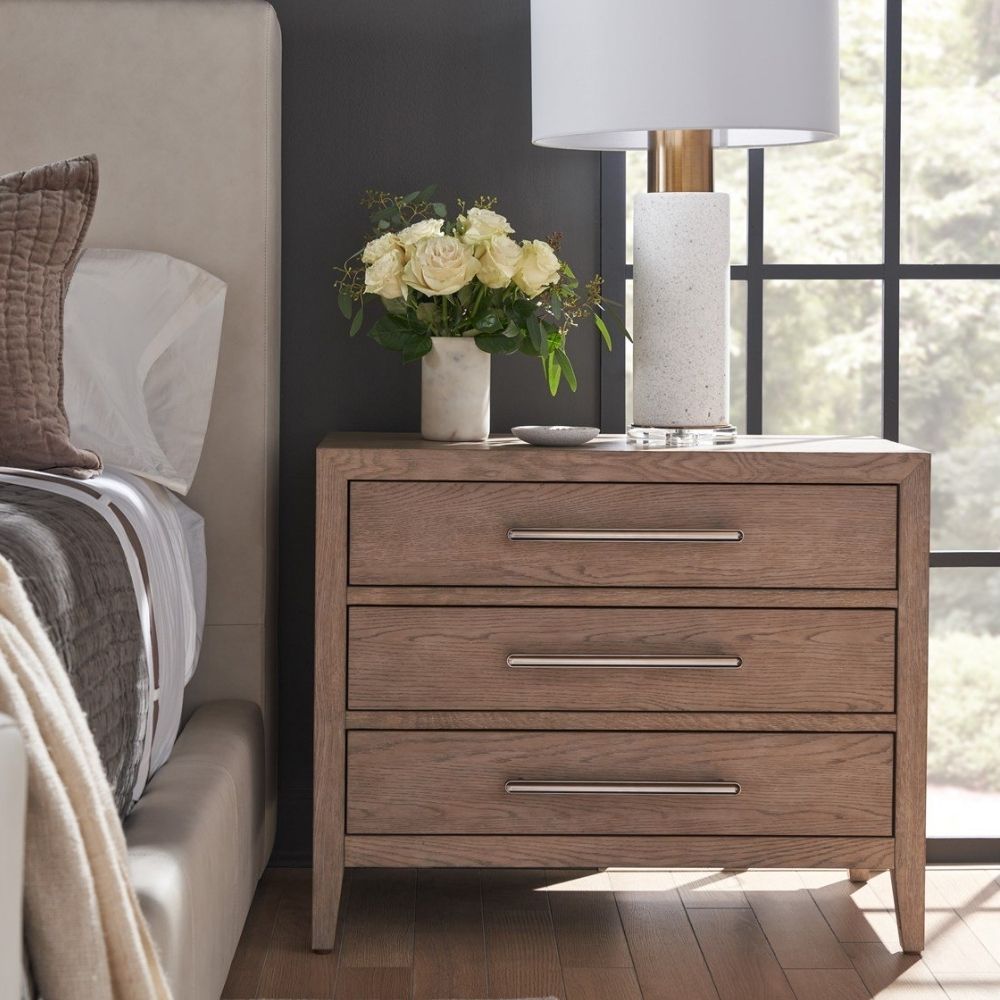 A three-drawer nightstand in a light natural wood finish with sleek horizontal metal handles and tapered legs.