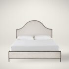 An upholstered bed in soft cream fabric with an arched headboard outlined by a slim dark metal frame, paired with a matching upholstered footboard.