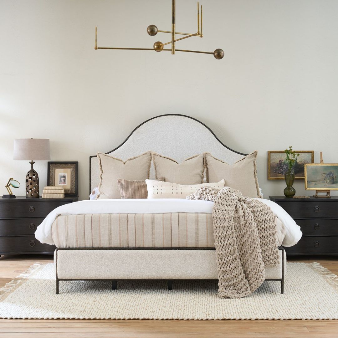 An upholstered bed in soft cream fabric with an arched headboard outlined by a slim dark metal frame, paired with a matching upholstered footboard.