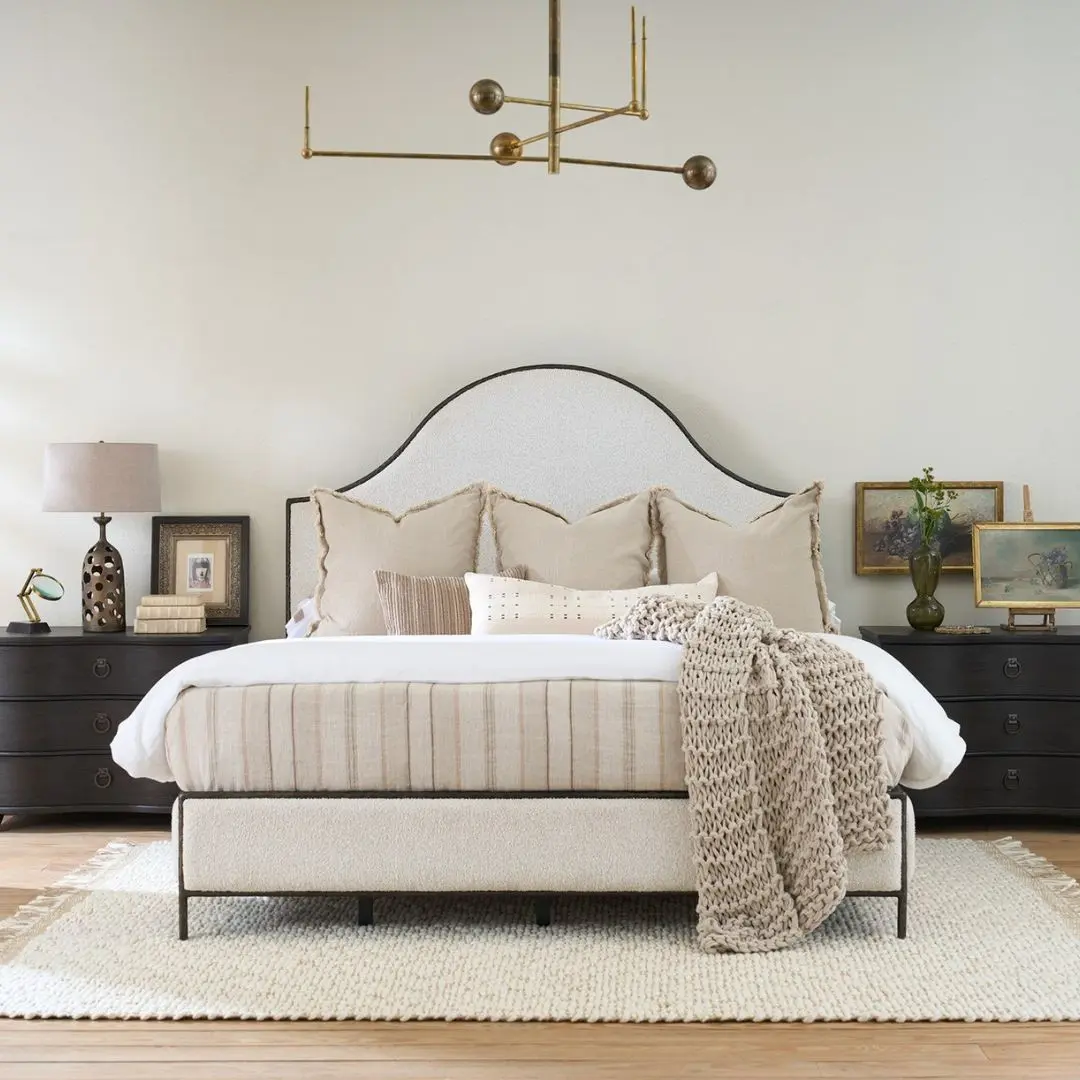 An upholstered bed in soft cream fabric with an arched headboard outlined by a slim dark metal frame, paired with a matching upholstered footboard.