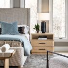A modern nightstand in a light natural wood finish with two drawers, a sleek silver handle design, and an open storage shelf.