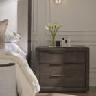 A dark wood nightstand with three drawers, slim black metal handles, and a bold plinth base for a modern grounded look.