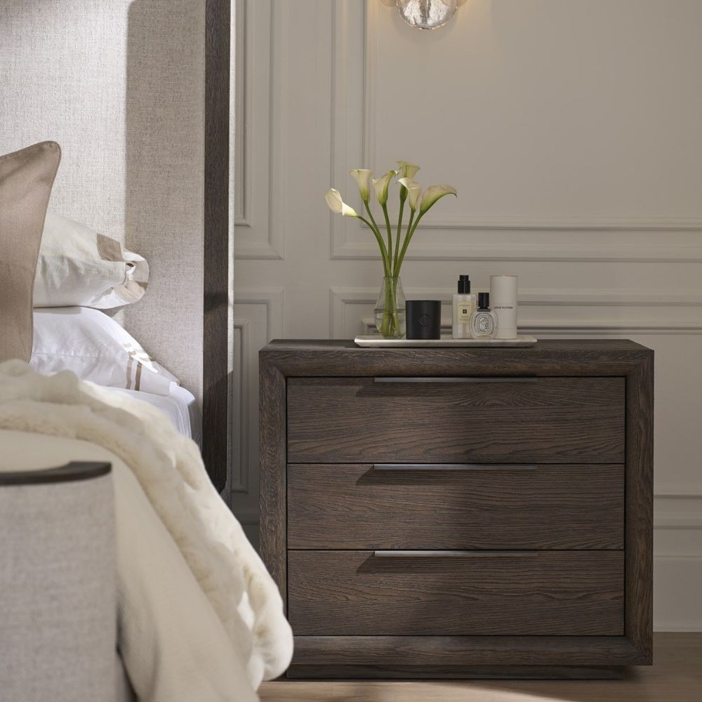 A dark wood nightstand with three drawers, slim black metal handles, and a bold plinth base for a modern grounded look.
