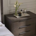 A dark wood nightstand with three drawers, slim black metal handles, and a bold plinth base for a modern grounded look.
